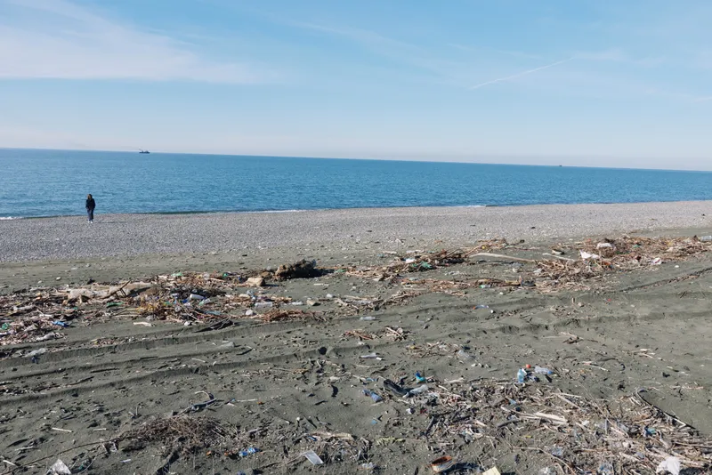 Solitary figure walks along a littered beach with debris and plastic waste, calm sea and clear horizon highlighting pollution and environmental impact.