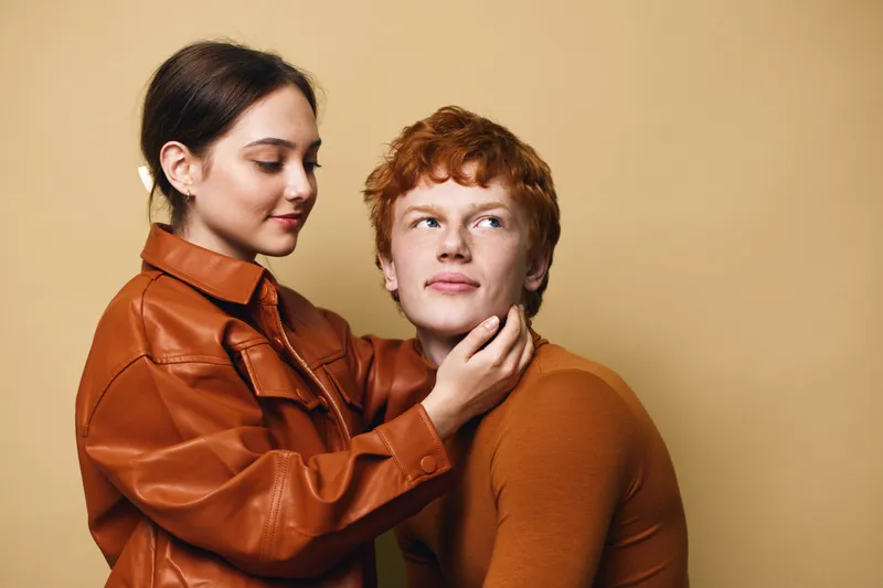 Two young people, woman and man, posing together in casual autumn clothes with warm colors, intimate and caring gesture with hand on face and gentle expression on beige background.