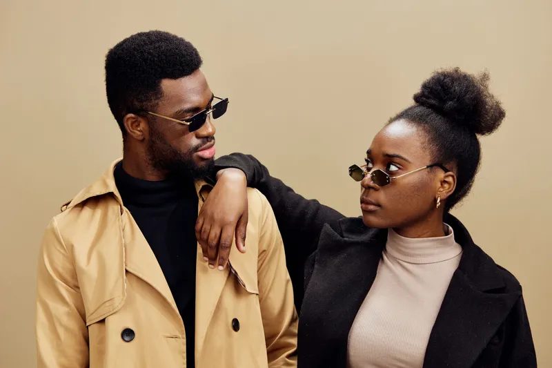 Stylish couple wearing sunglasses and fashionable outfits against a beige background, exuding confidence and contemporary style, showcasing modern fashion trends