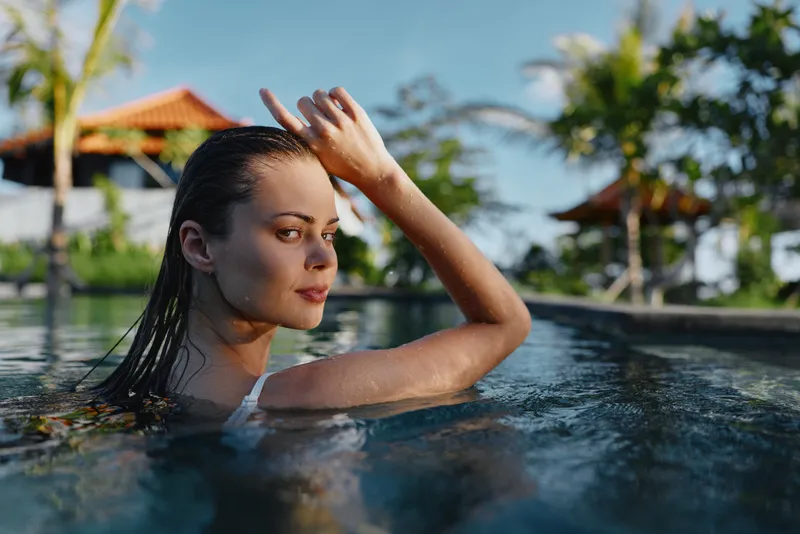Sunlit pool scene shows a woman partly submerged, brushing hair back with a calm gaze. Clear water, tropical backdrop, and relaxed mood convey leisure, wellness, and outdoor summer vibes.