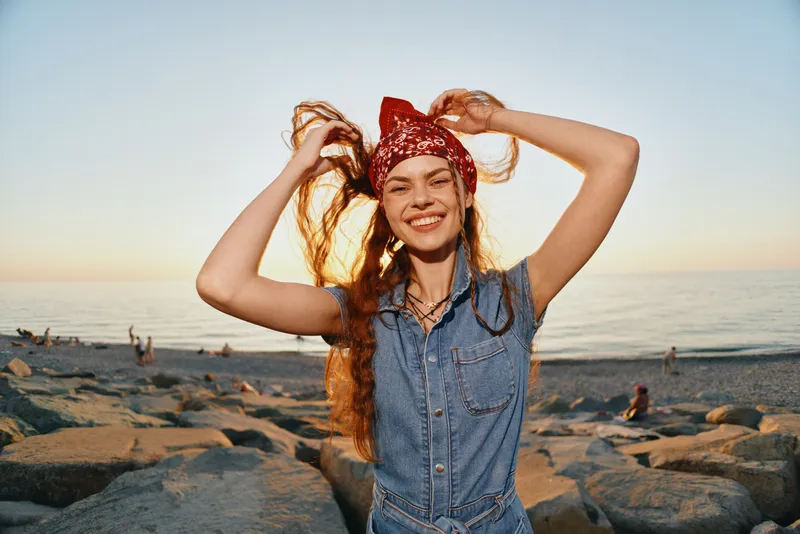 smiling woman with red bandana and denim dress enjoying sunny beach with golden light and ocean background in warm summer atmosphere