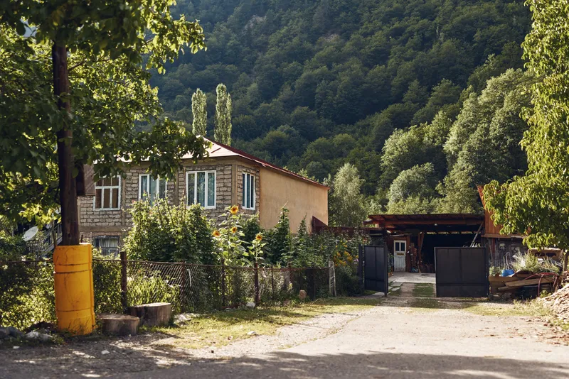 Rustic countryside house sits beside green hills with a gravel driveway, trees and a weathered facade, creating a peaceful rural scene bathed in natural light.