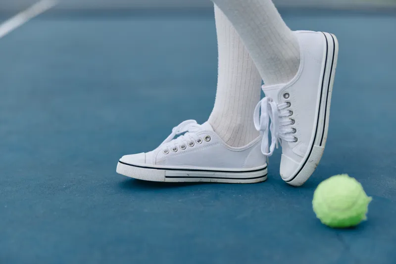 Young person in white sneakers and socks stands on a tennis court, ready to play with a green tennis ball, showcasing energy and anticipation