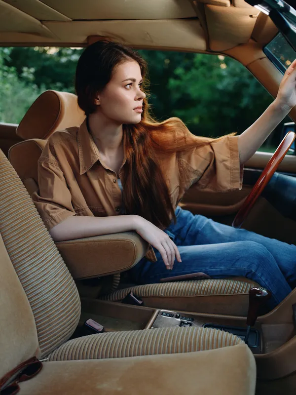 Young woman in casual attire seated in a vintage car, exuding a thoughtful expression, surrounded by lush green background, capturing a moment of nostalgia and freedom.