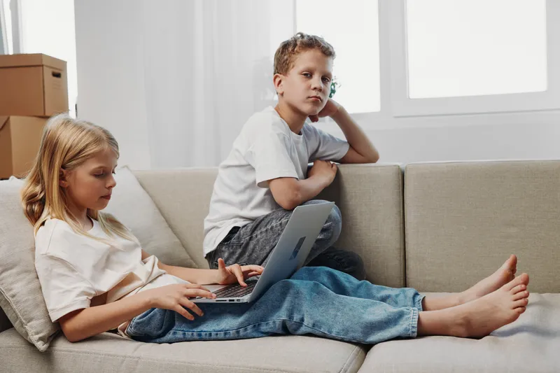 Two children using laptop on beige couch in bright room, girl focused on device and boy leaning on sofa armrest looking away. Concept of childhood, technology, and home environment.