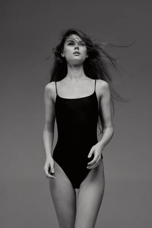 Black and white portrait of a young woman in a black bodysuit with windblown hair against a gray background.