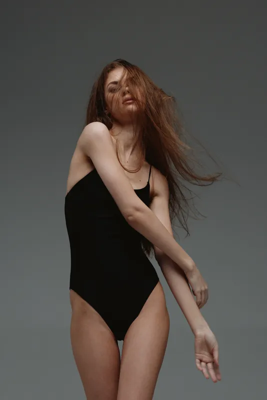 Slender woman with red hair wearing black bodysuit posing gracefully against gray background.