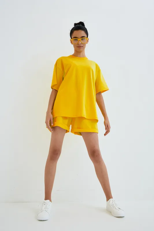 woman in bright yellow casual outfit standing confidently against white background, wearing matching shorts and oversized t-shirt with white sneakers