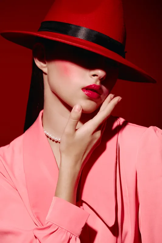 Fashion woman in red hat and pink jacket touches face showing glossy lips confinement. Creative beauty photography with bright makeup and pearl necklace on dark background. Concept fashion.