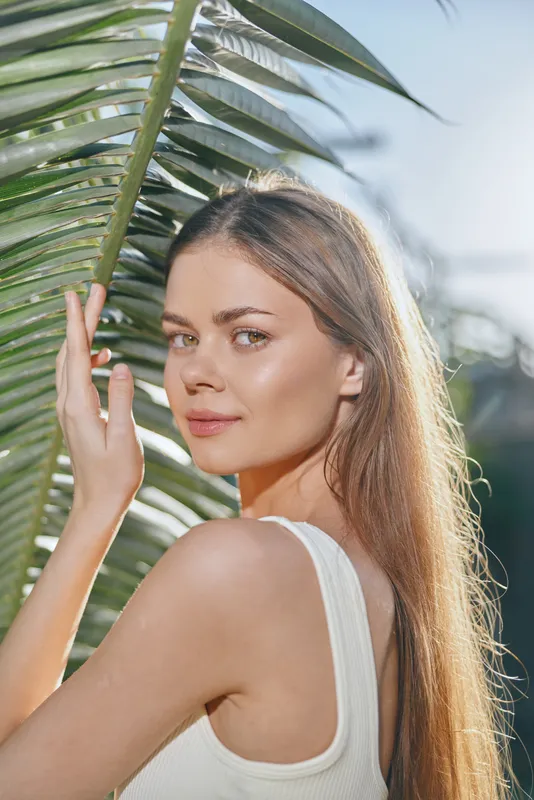 young woman posing near palm leaves outdoors with natural sunlight, long hair, gentle smile, summer vibes, casual white top, serene expression