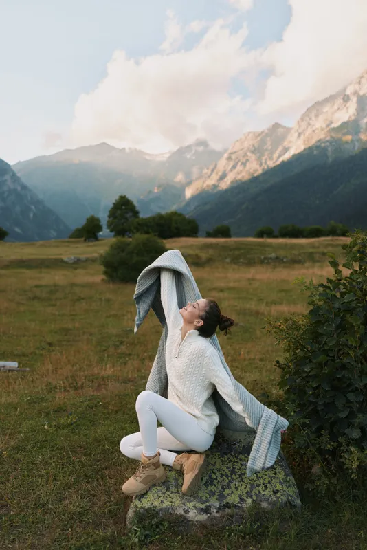 Young woman in white knitwear enjoys fresh mountain air while sitting on a rock and stretching with a scarf in an open field during a clear day.