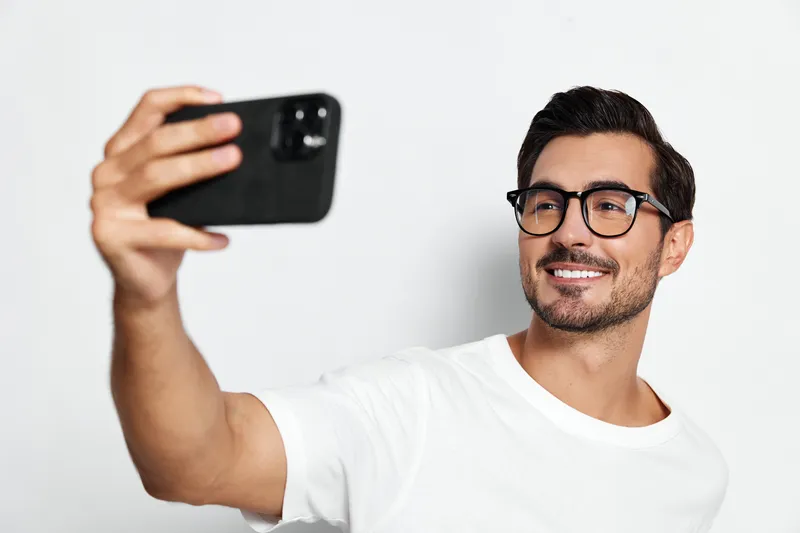 Smiling young man wearing glasses takes a selfie with smartphone, looking directly at the camera against a solid light background. Lifestyle concept.