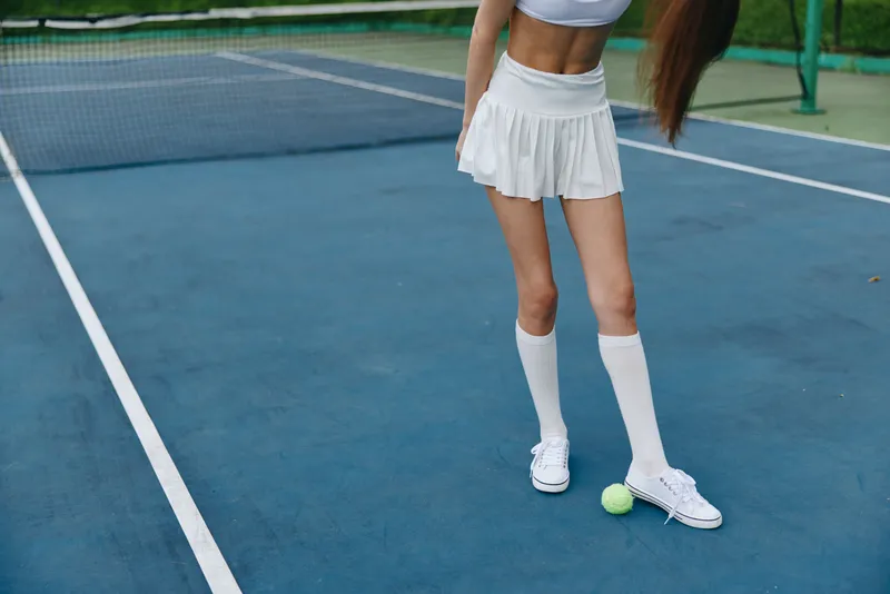 A young woman in a sporty white outfit poses on a tennis court, showcasing confidence and athleticism while standing near a tennis ball