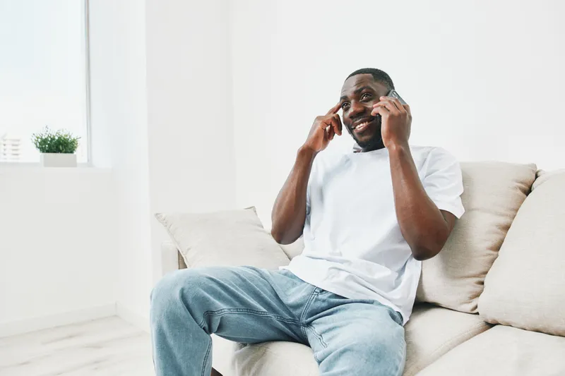 Young African American man smiling while talking on the phone at home, wearing casual clothing in a bright, minimalist interior