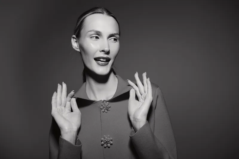 White woman with pale skin in vintage knitwear poses with hands raised, looking sideways. Studio portrait with soft lighting and classic mood.