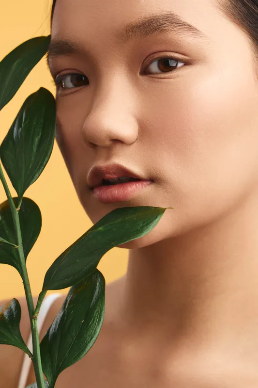 asian woman in a studio portrait gazes softly at the camera, with a leafy branch near her face, warm lighting and a minimal background creating a calm, natural mood.