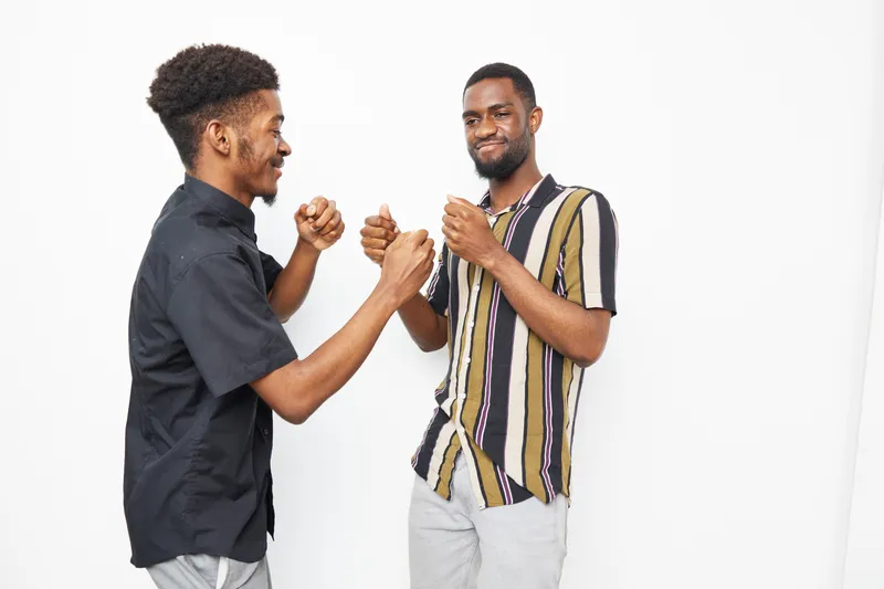 Two black men are playfully sparring, showcasing a lively mood. Their diverse outfits and expressions reflect friendship and energy in a bright, minimalist setting.