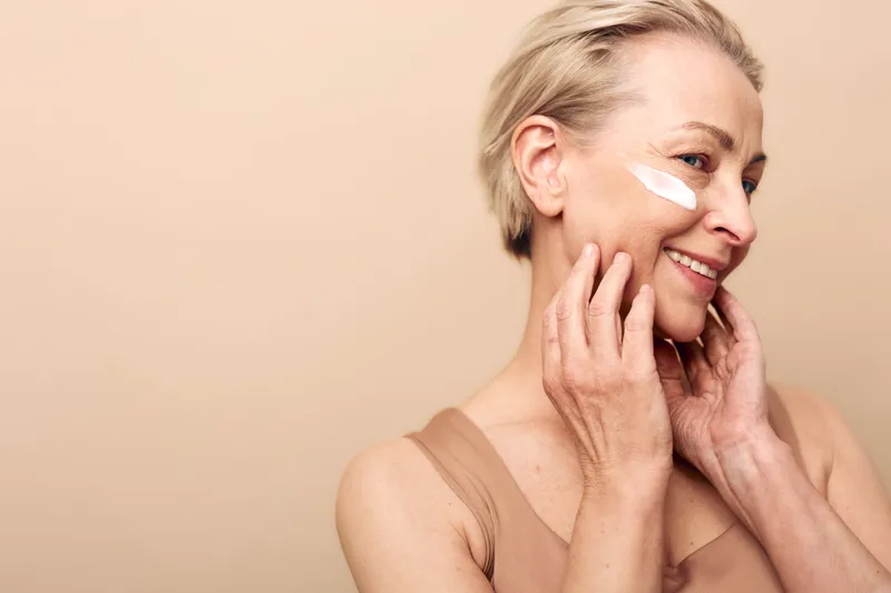 Smiling middle aged woman with short blonde hair applies white cream on cheek, touching face gently. Soft beige background and natural skincare concept with wellness and self care focus.