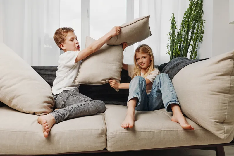 Two children having a playful pillow fight on a beige couch in a bright living room. Casual home environment shows fun, sibling interaction, and joyful childhood moments.