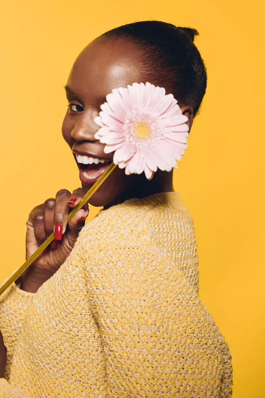 Happy dark skinned African woman in yellow sweater holds pink gerbera flower near face smiling on yellow background. Concept of joy, beauty, floral, and positive emotions.