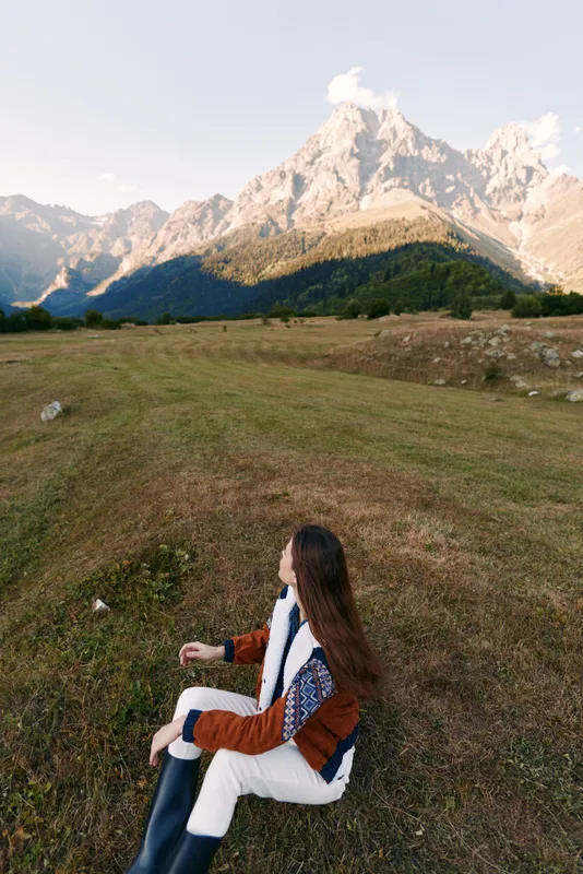 woman sitting on meadow facing mountain landscape in nature, autumn travel scene with long hair, cozy jacket and boots, peaceful outdoor relaxation, panoramic alpine valley and hiking vibe