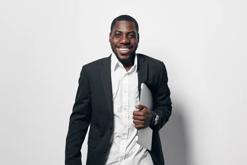 African American man with dark skin wearing black blazer and white shirt holding laptop smiling confidently standing against white background in studio portrait for business concept.