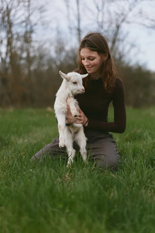 A woman sits on grass holding a small goat kid, sharing a calm moment outdoors. A lifestyle scene that highlights gentle affection and the bond between human and animal in a rural field.