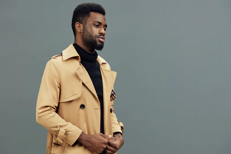 Stylish young African man in a beige trench coat against a minimalist gray background, exuding confidence and modern fashion