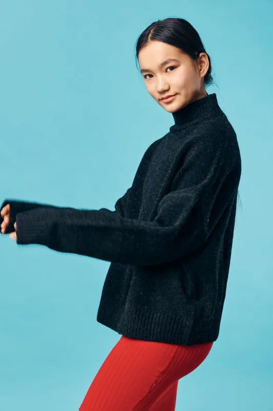 Young woman with black hair wearing black sweater and red pants posing against plain blue background. Casual style, studio portrait with confident expression and relaxed posture.