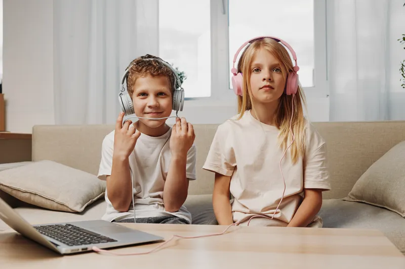 Two children wearing headphones sit on a couch with a laptop nearby, engaged in online learning or entertainment at home.