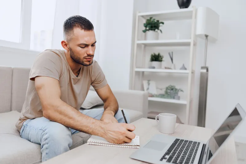 Focused man writing notes at home, surrounded by a modern and bright setting, showcasing productivity and creativity while working on a laptop.