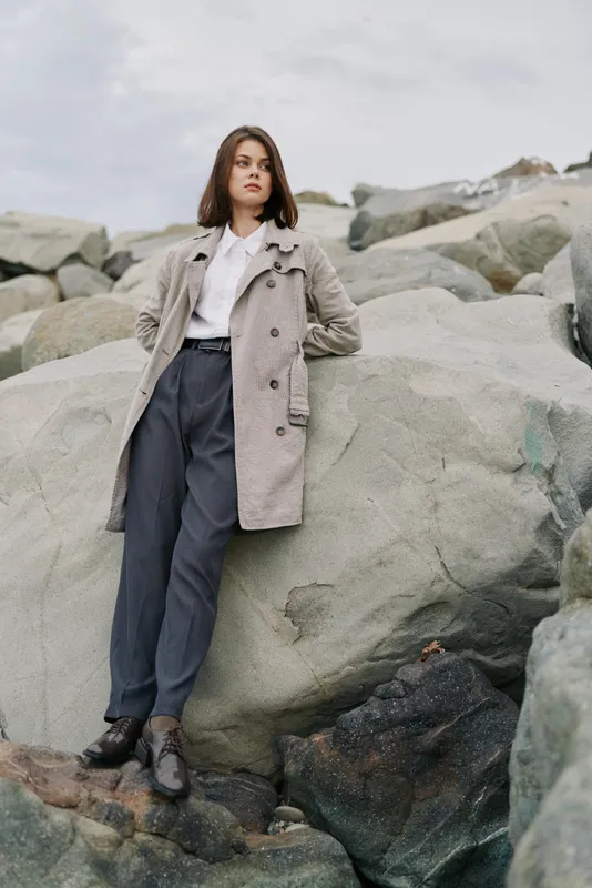 Young woman in stylish coat and dark trousers stands confidently on large rocks outdoors. Overcast sky and natural environment create a calm, contemplative mood in this fashion portrait.