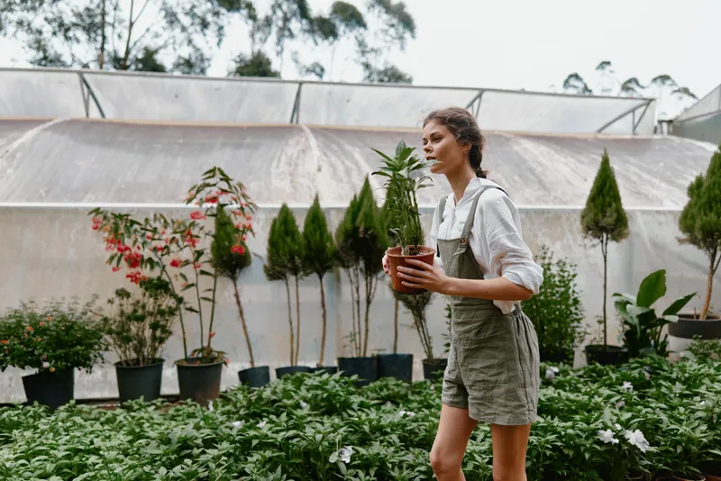 Young woman carrying a potted plant in a greenhouse, showcasing gardening passion, nature connection, and healthy lifestyle choices in a vibrant, seasonal setting