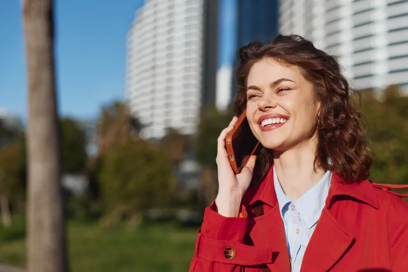 Young woman with curly hair wearing a red coat speaks on a smartphone in a sunny urban park, with modern buildings in the background and a bright, confident smile.