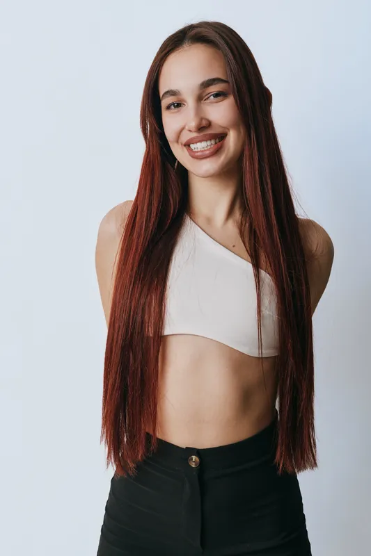 Young woman with long red hair smiling and posing against light background. Casual outfit with white crop top and black pants showing confidence and happiness in studio portrait.