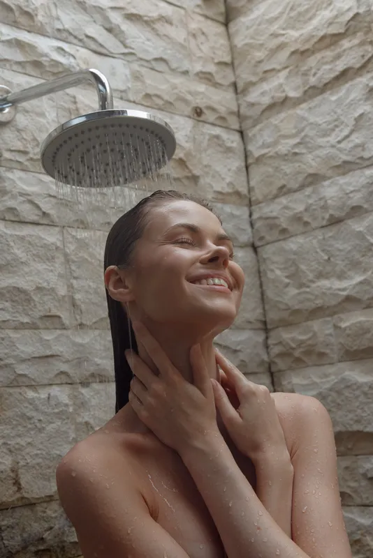 smiling woman enjoying outdoor shower with wet hair, natural stone wall background, serene expression, fresh skin, wellness concept, summer relaxation