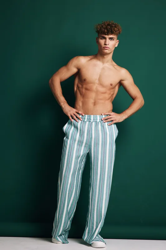 Young man posing shirtless with curly hair and striped pants standing with hands on hips against green background. Confident model demonstrating fit physique and fashion style.