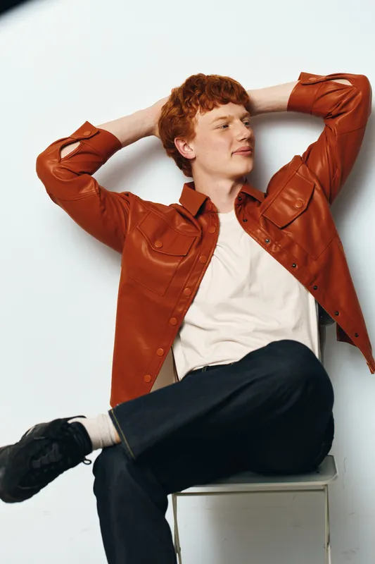 Young man with red hair in brown shirt and white t shirt sits relaxed on chair with crossed legs against white background in casual pose.
