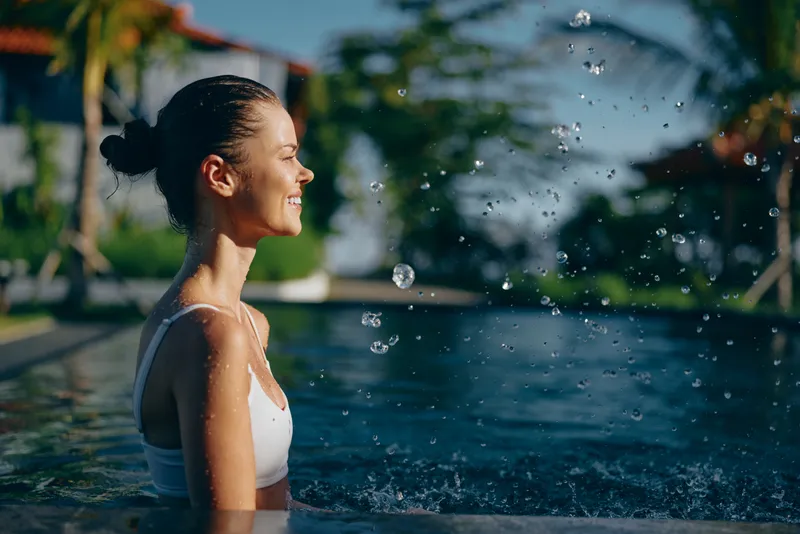 Cheerful woman in a white top stands in an outdoor pool, smiling with water droplets in the air, sunlit scene, lush greenery background, relaxed mood, candid summer leisure moment.