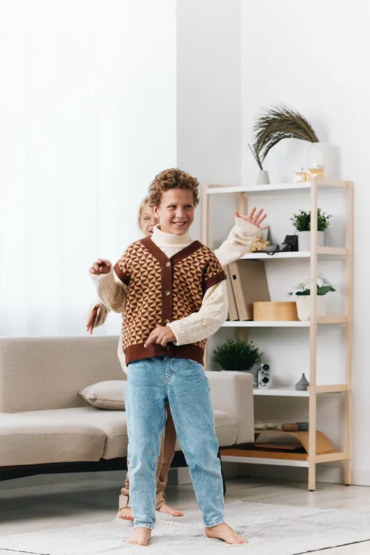 Playful child dances indoors wearing a cozy knit vest and layered sweater, smiling and full of energy as they move across a bright living room with soft decor and plush rug.