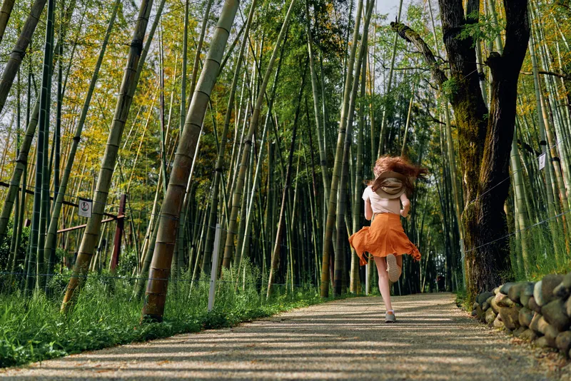 Girl running through a bamboo forest on a gravel path, back view in orange skirt and dress, motion and sunlight through canopy, outdoor nature scene for travel and freedom.
