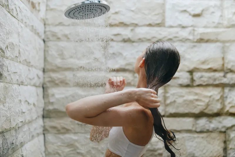 Woman taking outdoor shower with flowing water on stone wall background, relaxed young adult enjoying refreshing moment in natural setting during daytime.