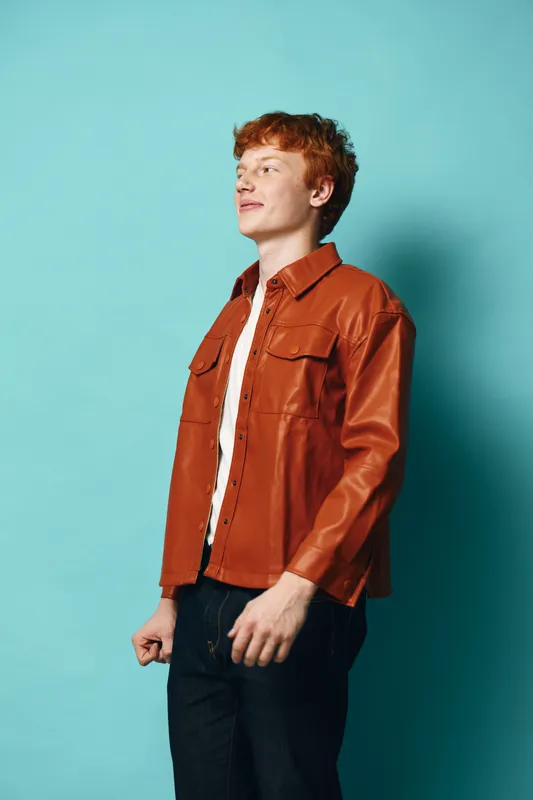 Young man with red hair wearing brown leather jacket and white shirt standing against blue background looking sideways with calm expression and casual pose.