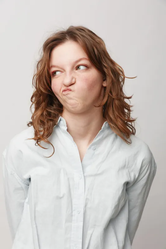 Young woman with curly hair wearing white shirt makes a disgusted face looking sideways. Isolated on light background. Concept of dislike, confusion, and negative emotion.