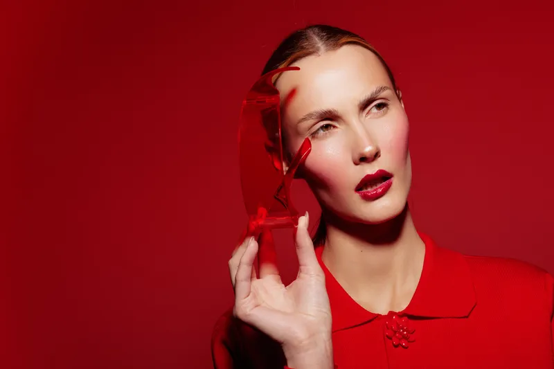 Woman with red lipstick and red blouse holds red glass object near face on red background. Concept of fashion, beauty, and artistic portrait with monochrome color scheme and creative styling.