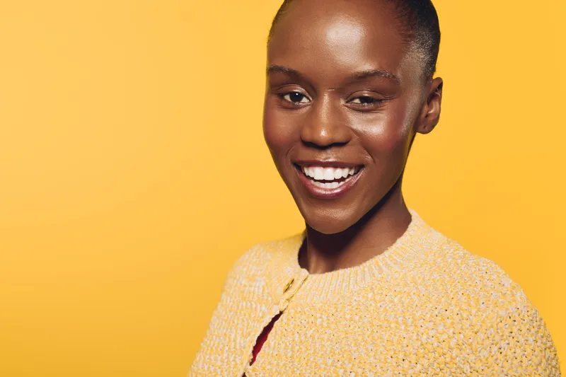 Smiling African American woman with dark skin and short hair poses against yellow background wearing cozy sweater. Concept of happiness, beauty, confidence, and natural look in studio lighting.