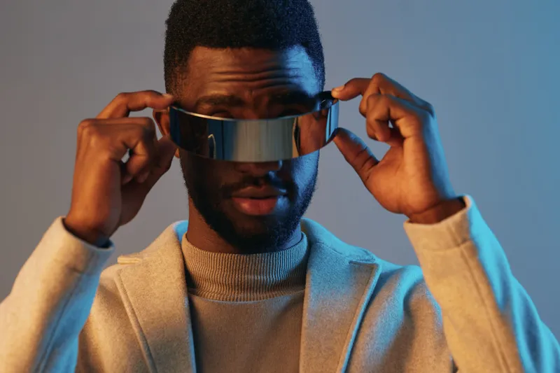 A stylish African man in futuristic sunglasses confidently poses against a blue backdrop, showcasing contemporary fashion and bold expressions, perfect for modern lifestyle concepts