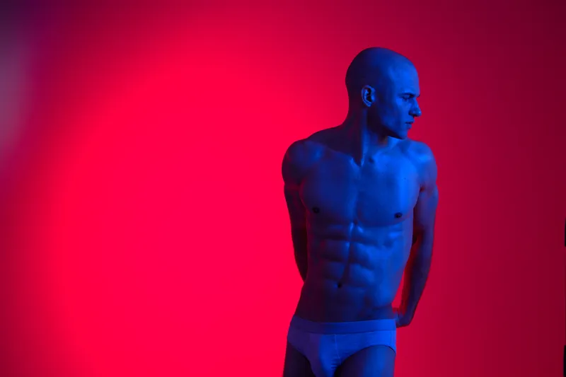 Muscular man posing against a vibrant background, showcasing strength and confidence. The light creates a dramatic effect, highlighting his toned physique.