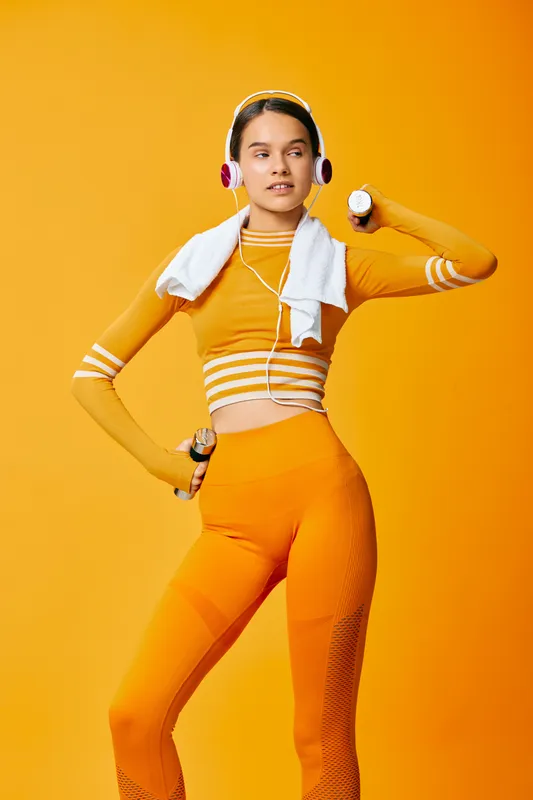 Fit woman in orange activewear holding dumbbells and wearing headphones with a towel over her shoulders, striking a confident pose against a vibrant orange studio background.