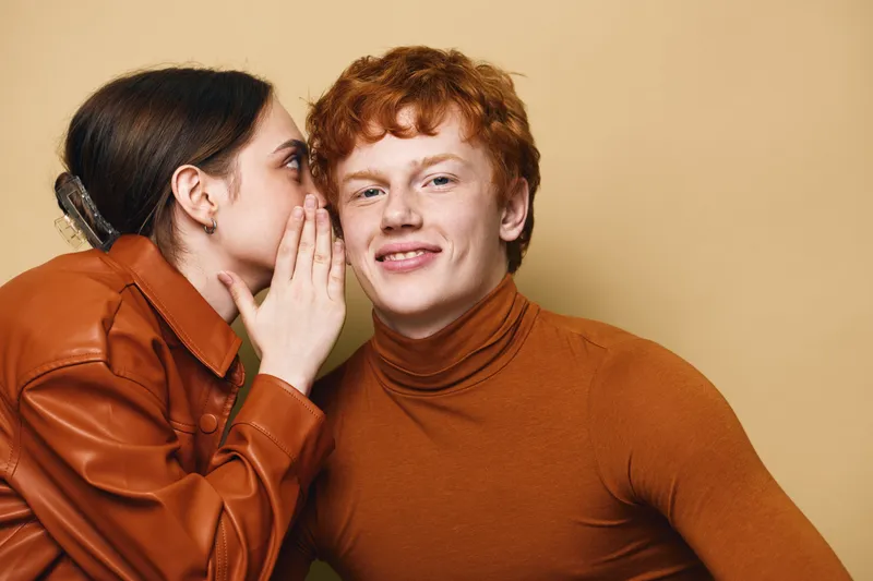 Young woman whispering to young man with red hair in casual clothing for secret communication and friendship indoor. Closeup, studio, human expression and communication between two friends.